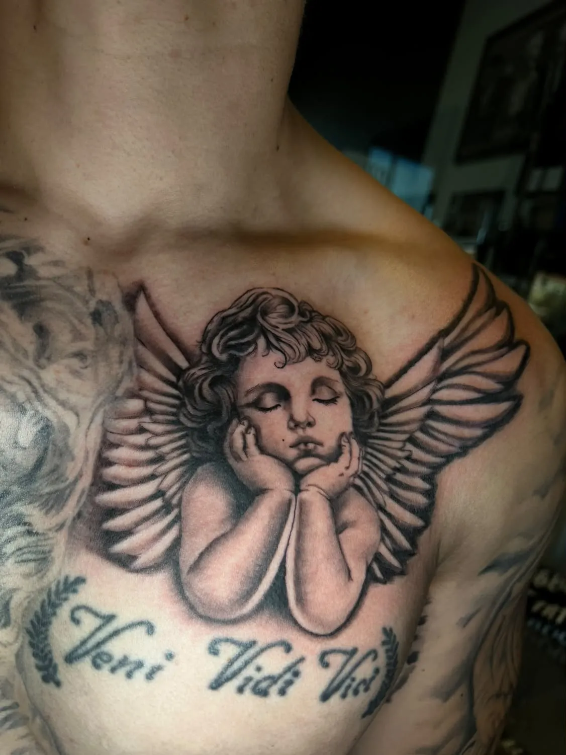 Realistic black-and-gray cherub with closed eyes and open wings, resting its chin on its hands across the upper chest