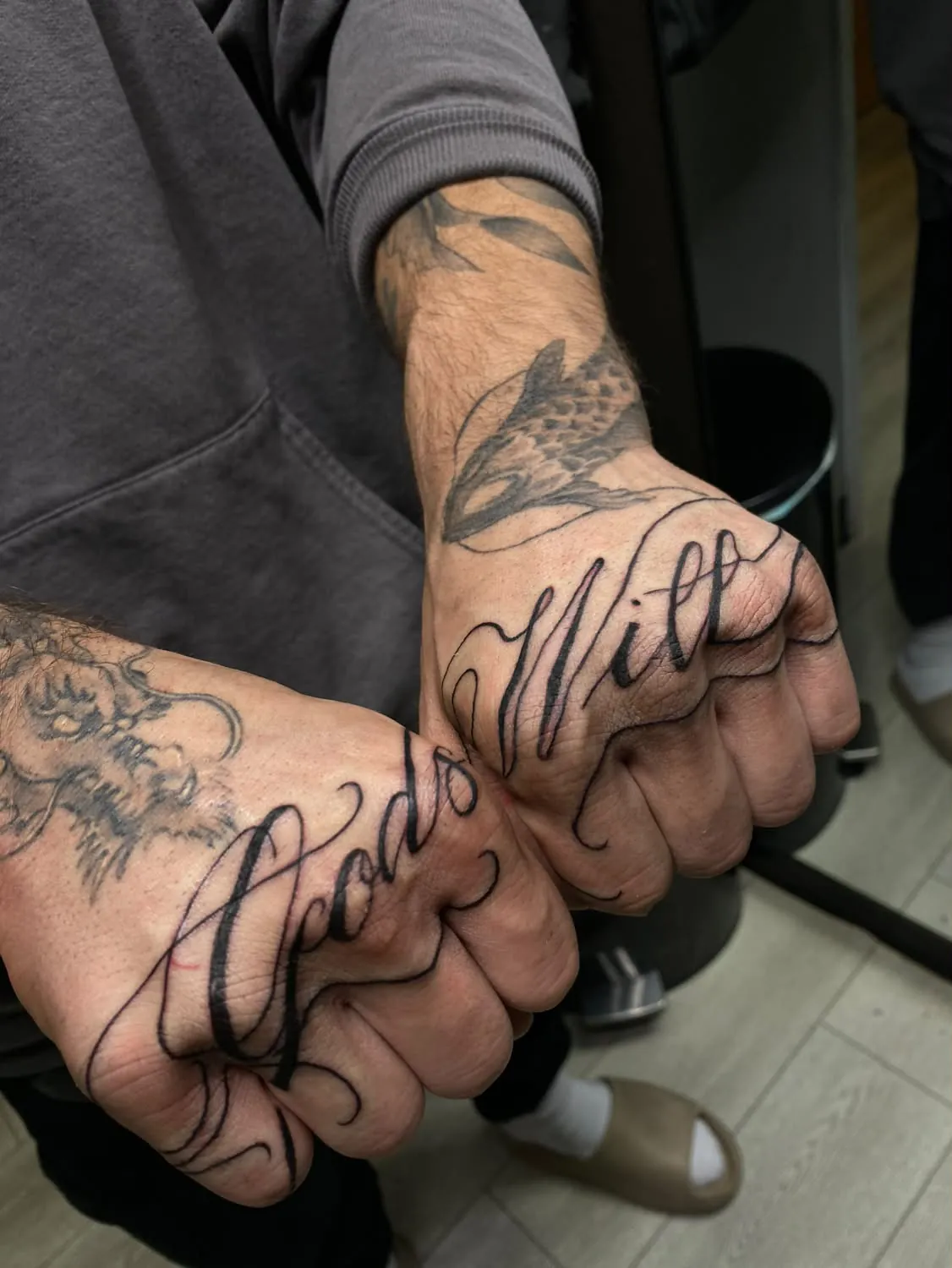 Flowing script lettering reading “Gods Will” spread across the backs of two clenched fists